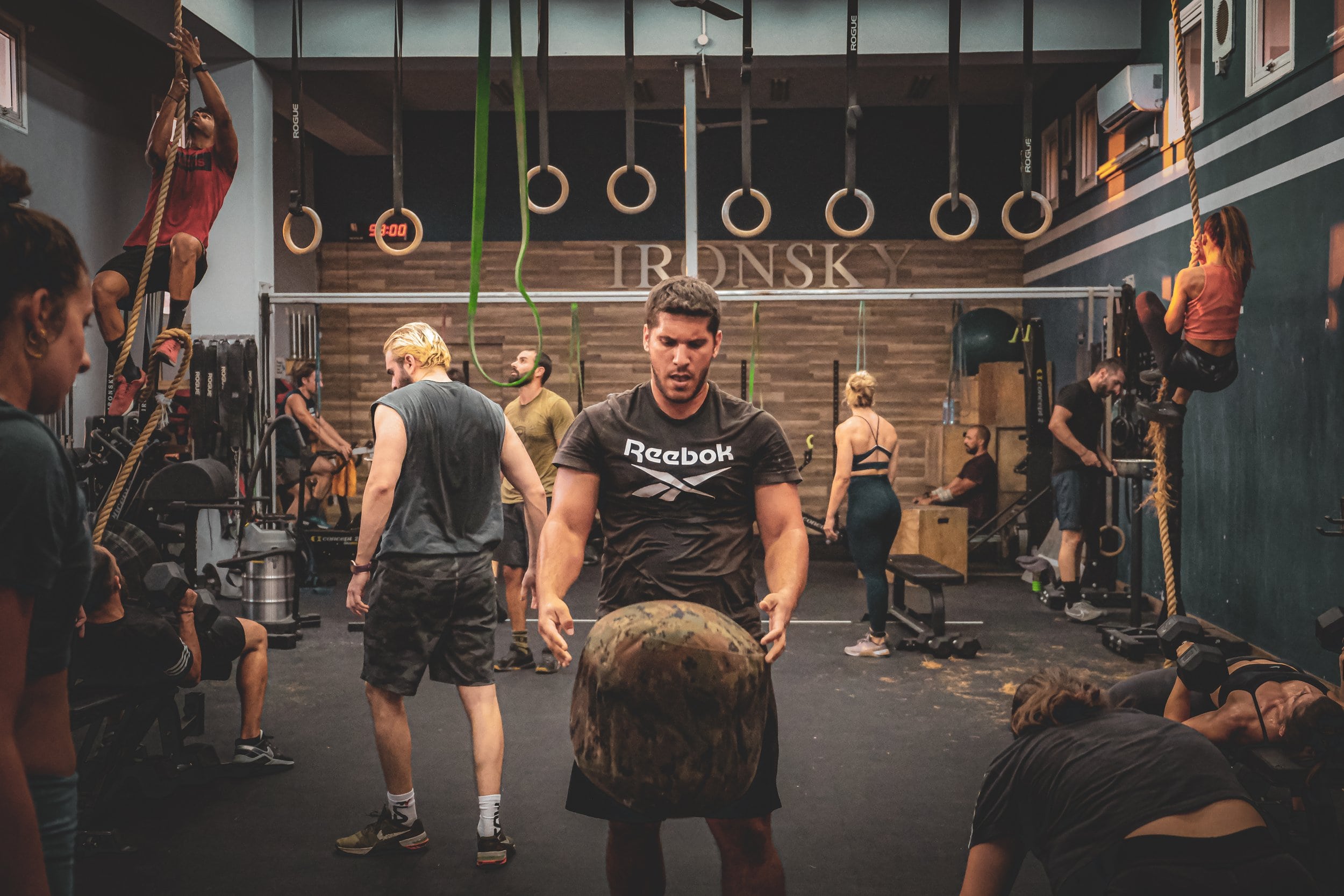 A group of people engaged in various workout activities at a well-equipped gym, including rope climbing, weightlifting, and using fitness machines.