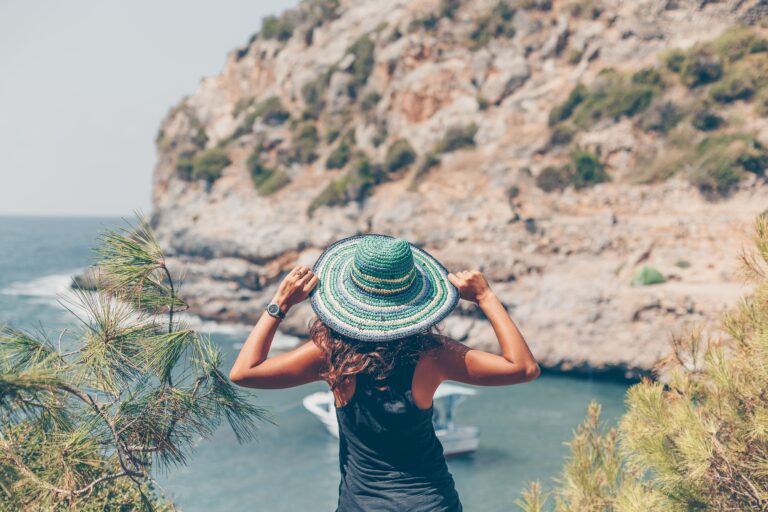 Woman wearing a colorful straw hat overlooking a coastal view with rocky cliffs and a boat on the water in Cyprus.