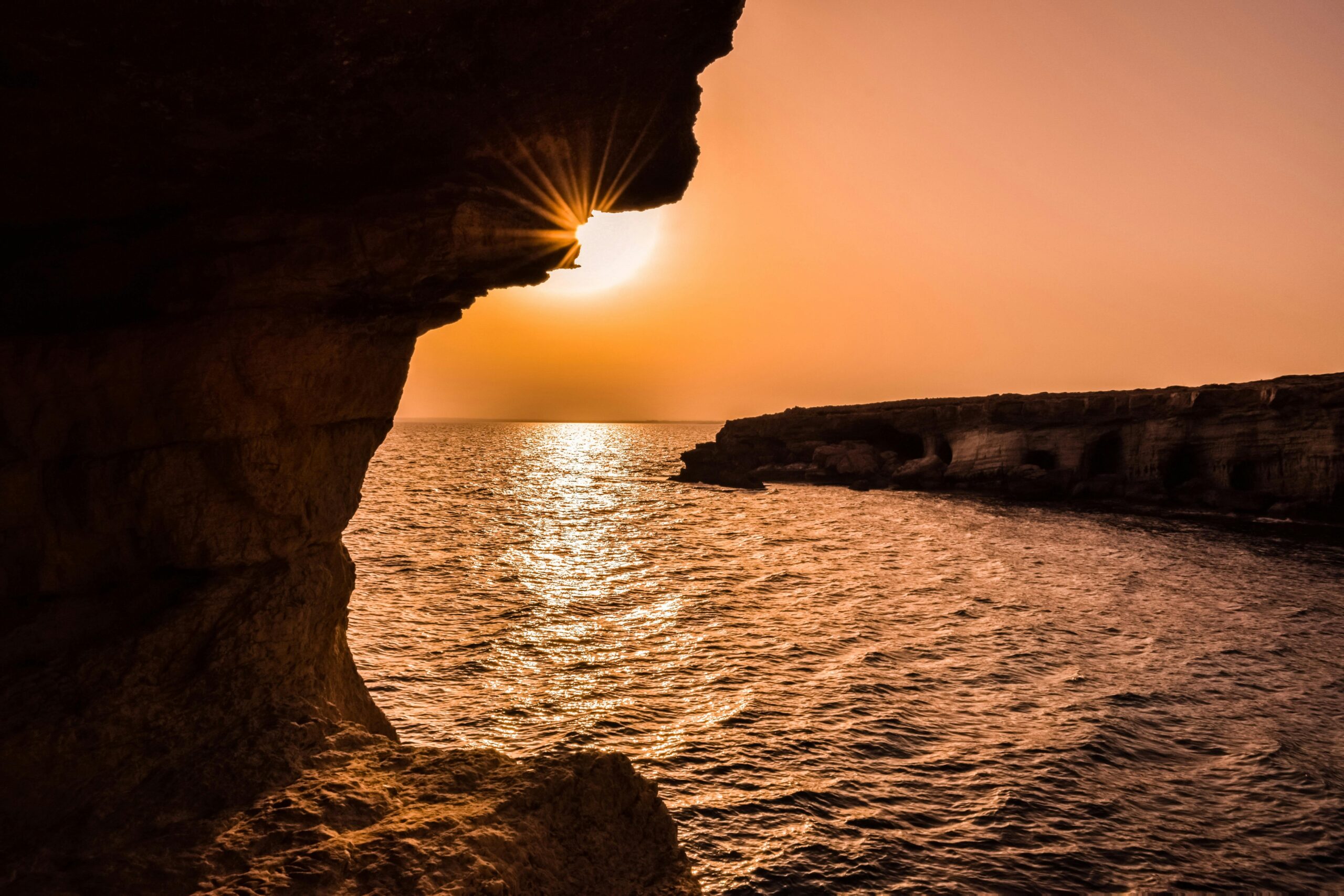 Sunset over the sea with dramatic cliffs and the sun peeking through a rock formation in Cyprus.