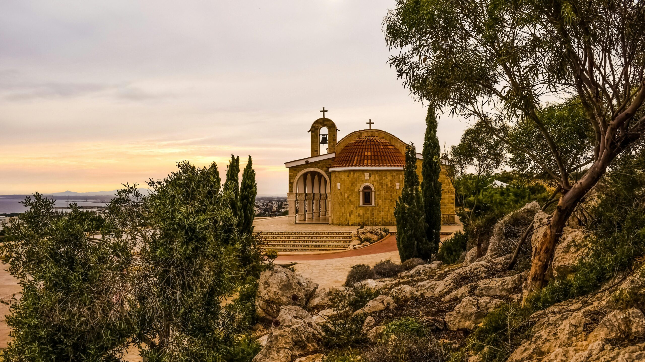 A charming small chapel with a red-tiled roof and bell tower, surrounded by trees and rocky terrain in Cyprus during sunset.