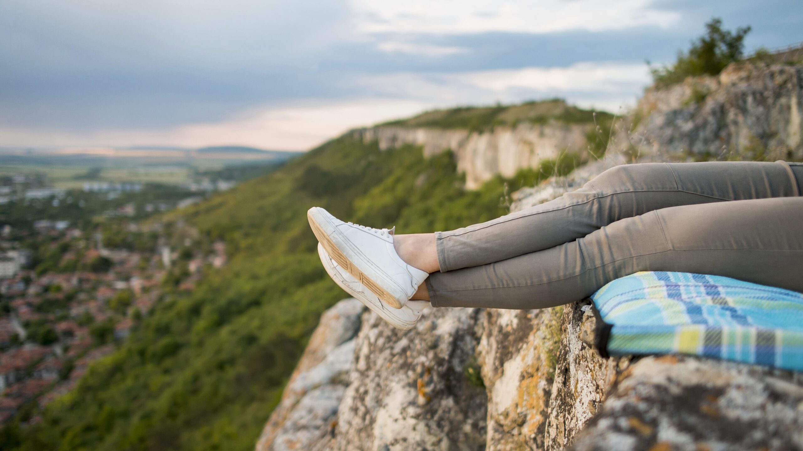 Person relaxing on a cliffside overlooking a scenic valley, legs extended with white sneakers visible.