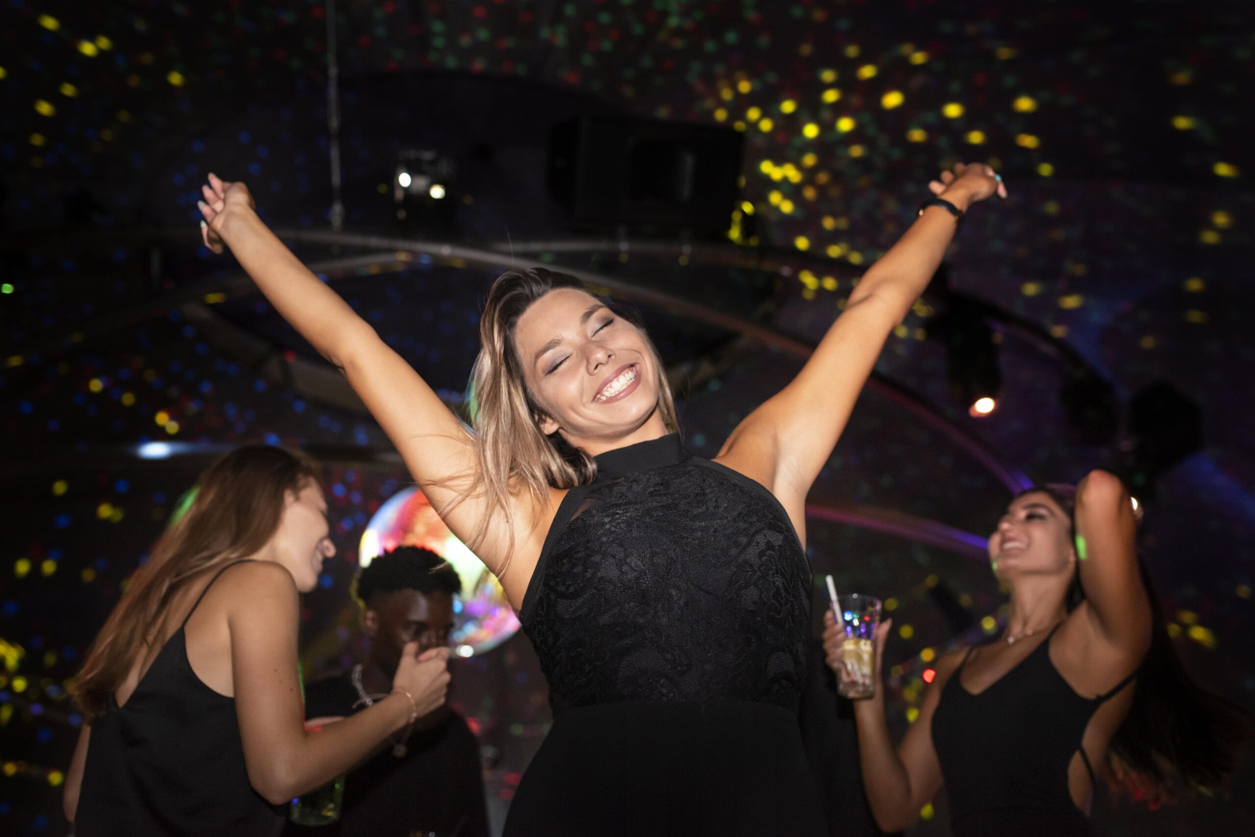 A smiling woman with arms raised dancing at a nightclub, surrounded by friends and colorful lights.