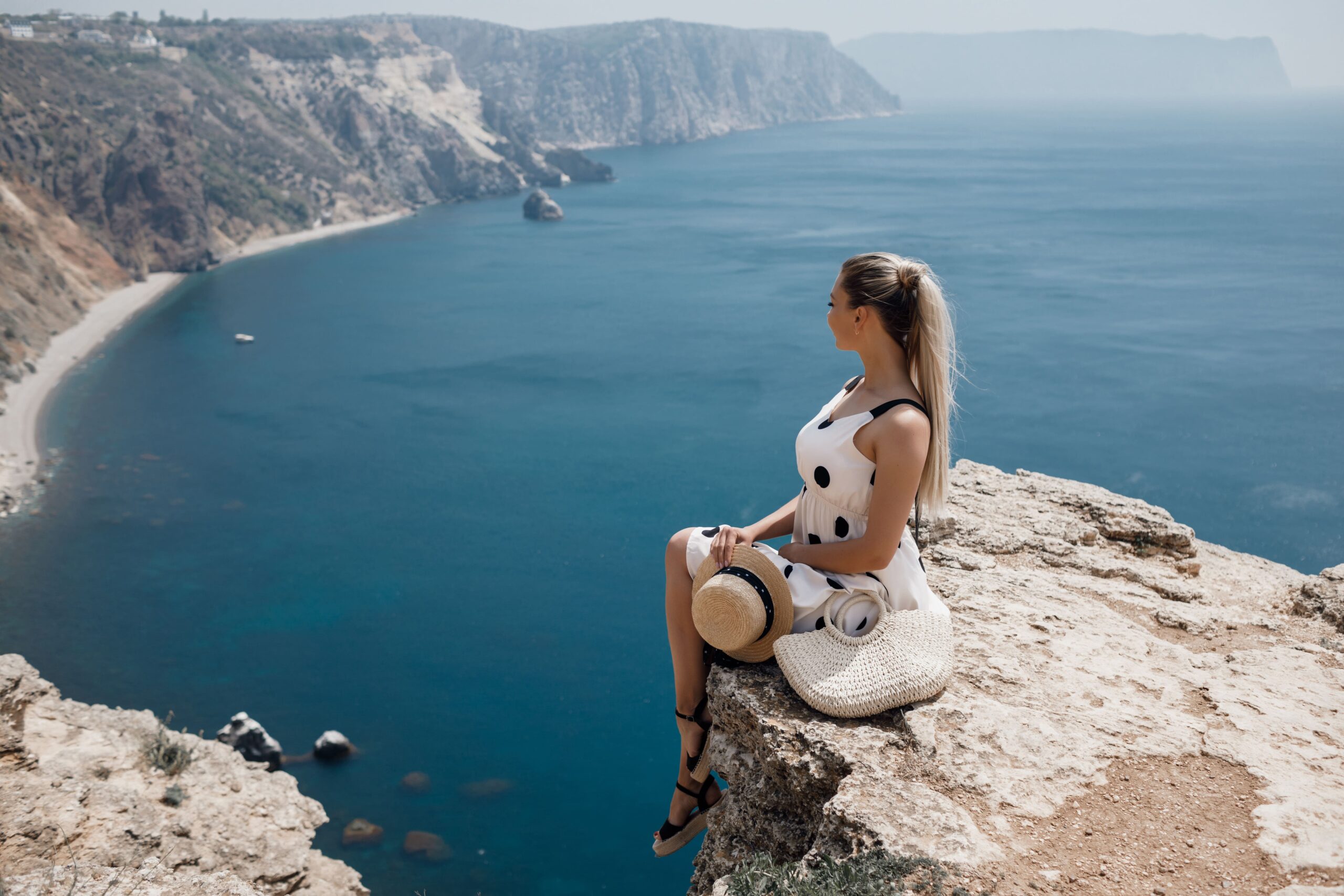 Woman wearing a colorful straw hat overlooking a coastal view with rocky cliffs and a boat on the water in Cyprus.